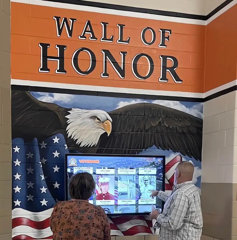 Two visitors explore a Wall of Honor touchscreen beneath a painted eagle and flag